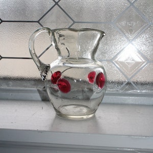 Puede incluir: Una jarra de vidrio transparente con un asa plateada y tres cerezas rojas en el lateral.