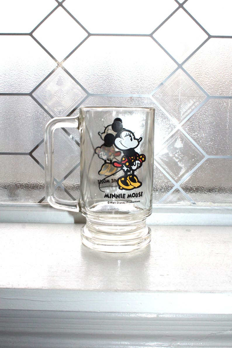 May include: A clear glass mug featuring a graphic of Minnie Mouse in a red and yellow polka dot dress with yellow shoes. The text MINNIE MOUSE is printed in black below the character, with the text Walt Disney Productions printed underneath.