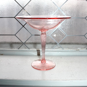 May include: A pink glass dessert dish with a wide, shallow bowl and a twisted stem. The rim of the bowl has a darker pink accent. The dish is displayed on a white windowsill in front of a window with a diamond pattern.
