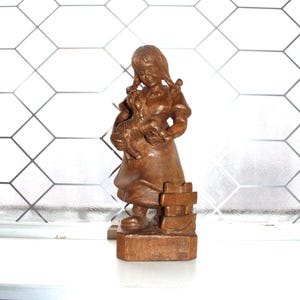 以下が含まれることがあります： 犬を抱きかかえる少女の木製フィギュア。少女はドレスを着て、小さな木の柵の隣に立っています。茶色で、詳細な彫刻が施されています。