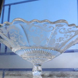 May include: A clear glass bowl with a scalloped edge and an etched floral design. The bowl is on a pedestal stand.