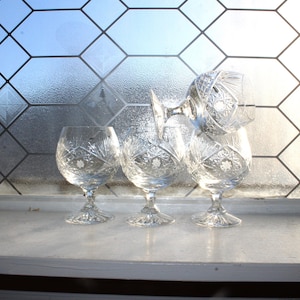 May include: Four clear crystal brandy snifters with intricate starburst and floral patterns. The glasses are arranged on a white windowsill in front of a window with a geometric design. The glasses are illuminated by natural light.