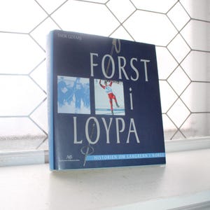 May include: A hardcover book titled "FORST i LOYPA" by Thor Gotaas. The cover is navy blue with white text and two images. One image shows a group of people, and the other shows a person skiing. The book is on a white windowsill.