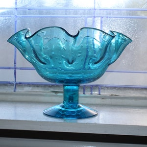 May include: A blue glass bowl with a ruffled rim and a pedestal base. The bowl has a textured surface with small bubbles.