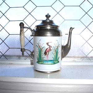 May include: A white and silver teapot with a decorative handle and spout. The teapot features a painted scene of a pink heron standing in water with reeds. The lid and trim are silver. The handle is cream-colored with a decorative design.