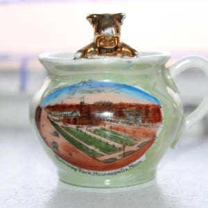 May include: A small, green ceramic mug with a white handle and a gold-colored dog figurine on top. The mug features a painted scene of Gateway Park, Minneapolis, Minnesota.