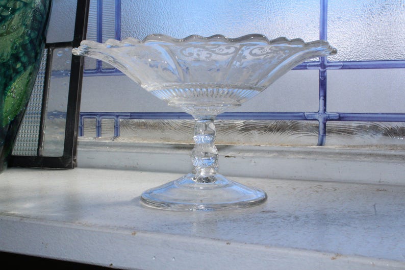 May include: A clear glass pedestal bowl with a scalloped edge and intricate etched designs. The bowl is on a white surface.