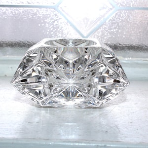 May include: A clear, faceted crystal votive candle holder. The geometric design features diamond-shaped cuts that reflect light. The candle holder is sitting on a white surface, with a window in the background.