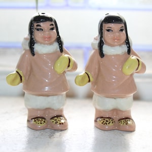 Könnte beinhalten: Ein Paar Salz- und Pfefferstreuer aus Keramik in Form von Personen, die traditionelle Winterkleidung tragen. Die Streuer sind in den Farben Rosa, Weiß und Gelb bemalt.