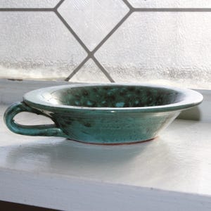 Op de afbeelding: Een kleine, groene keramische kom met een gespikkeld glazuur en een handvat. De kom staat op een wit oppervlak.