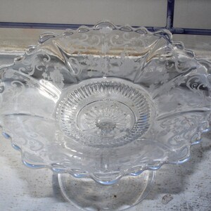 May include: A clear glass pedestal bowl with an intricate etched floral design. The bowl has a scalloped edge and a raised center.