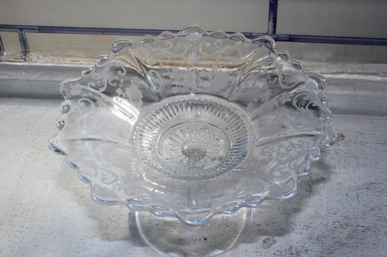 May include: A clear glass pedestal bowl with an intricate etched floral design. The bowl has a scalloped edge and a raised center.