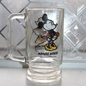 May include: A clear glass mug featuring a graphic of Minnie Mouse wearing a red and yellow polka dot dress and yellow shoes. The text MINNIE MOUSE is printed in black below the character, with the text Walt Disney Productions underneath it.