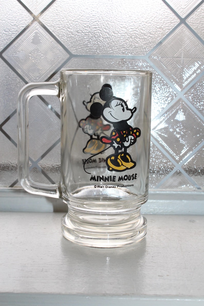 May include: A clear glass mug featuring a graphic of Minnie Mouse wearing a red and yellow polka dot dress and yellow shoes. The text MINNIE MOUSE is printed in black below the character, with the text Walt Disney Productions underneath it.