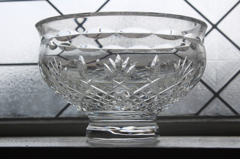Puede incluir: Un cuenco de cristal tallado transparente, de forma ancha y redondeada. El cuenco presenta intrincados patrones de diamantes y hojas, con una base gruesa y un borde liso. El cristal refleja la luz, creando un efecto brillante. Ideal para servir o decorar.