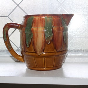 May include: A brown ceramic pitcher with a green and brown glaze. The pitcher is shaped like a barrel and has a handle.