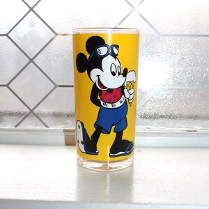 May include: A clear glass tumbler featuring a bright yellow background with a graphic of Mickey Mouse wearing blue sunglasses, a red polka-dot shirt, and blue shorts while holding an ice cream cone. The character is depicted in black and white.