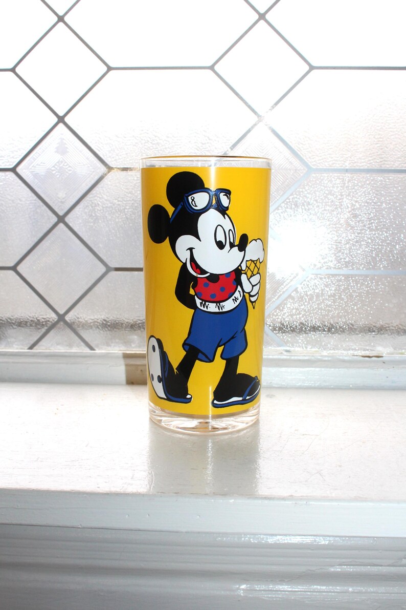 May include: A clear glass tumbler featuring a bright yellow background with a graphic of Mickey Mouse wearing blue sunglasses, a red polka-dot shirt, and blue shorts while holding an ice cream cone. The character is depicted in black and white.