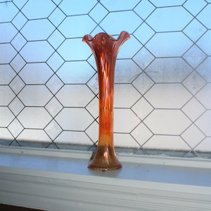 May include: A tall, amber-colored glass vase with a flared top and a ribbed design. The vase is sitting on a white windowsill in front of a window with a diamond-shaped grid pattern.