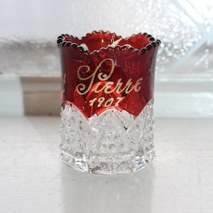 May include: A vintage red and clear glass votive holder. The top half is a deep red with the name "Pierre" and the year "1901" in gold script. The bottom half is clear, cut glass with a diamond pattern.