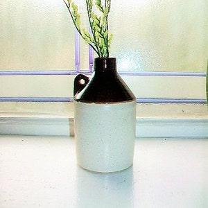 May include: A small, white ceramic pitcher with a brown top and a handle. The pitcher is filled with a bouquet of light green flowers.