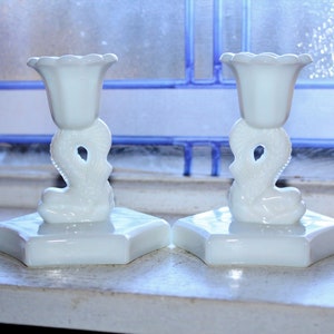 May include: Two white ceramic candle holders with a floral design on the top and a decorative base. The holders are on a white surface.