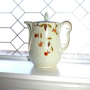 May include: Cream-colored ceramic pitcher with a lid, adorned with a floral pattern in red, yellow, and brown. The pitcher has a curved handle and gold trim, sitting on a white windowsill. The background features a window with a diamond grid.