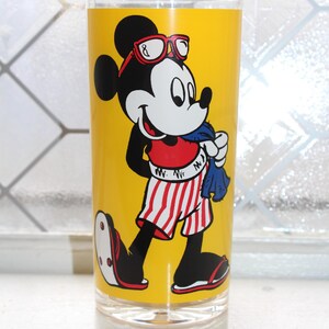 May include: A clear glass tumbler featuring a vibrant yellow background with a graphic of Mickey Mouse wearing red-rimmed glasses, a red shirt, and red-and-white striped shorts. Mickey is holding a blue towel and wearing sandals on his feet.