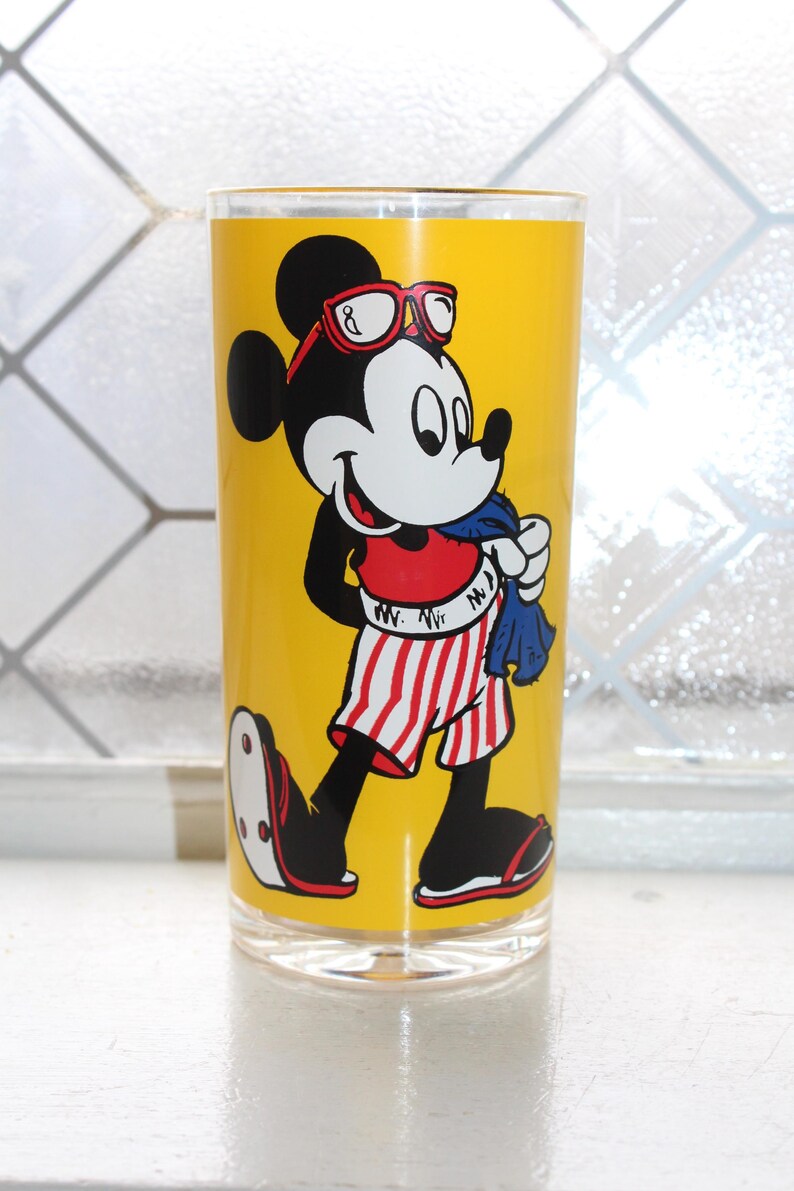 May include: A clear glass tumbler featuring a vibrant yellow background with a graphic of Mickey Mouse wearing red-rimmed glasses, a red shirt, and red-and-white striped shorts. Mickey is holding a blue towel and wearing sandals on his feet.