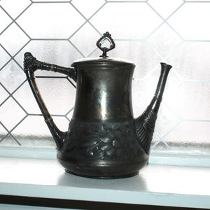 May include: A silver teapot with an ornate design and a decorative handle. The teapot has a floral pattern and is sitting on a white windowsill.