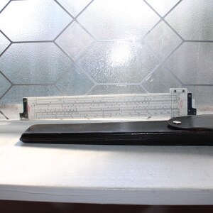 May include: A white plastic slide rule with black numerical markings and a sliding cursor, positioned behind a dark brown leather protective case with a silver-colored metal snap closure. The device features various scales and red text markings.