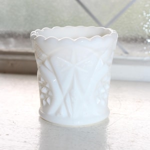 May include: A white milk glass votive holder featuring a scalloped top edge and a textured geometric pattern of raised stars and diamond shapes around the exterior. The cylindrical vessel has a smooth base and a translucent, opaque finish.