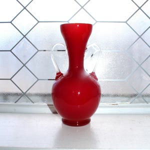 May include: A vibrant red glass vase with a bulbous body and two clear, curved handles. The vase has a flared top and sits on a small, circular base. The background features a window with a diamond-patterned design.