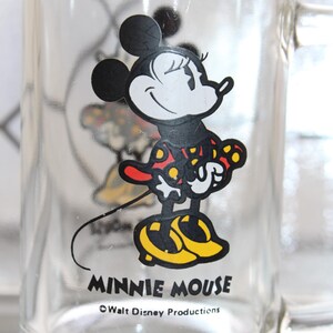 May include: A clear glass mug featuring a graphic of Minnie Mouse wearing a red dress with yellow polka dots and yellow shoes. The text MINNIE MOUSE is printed in black below the character, followed by the text &copy;Walt Disney Productions.