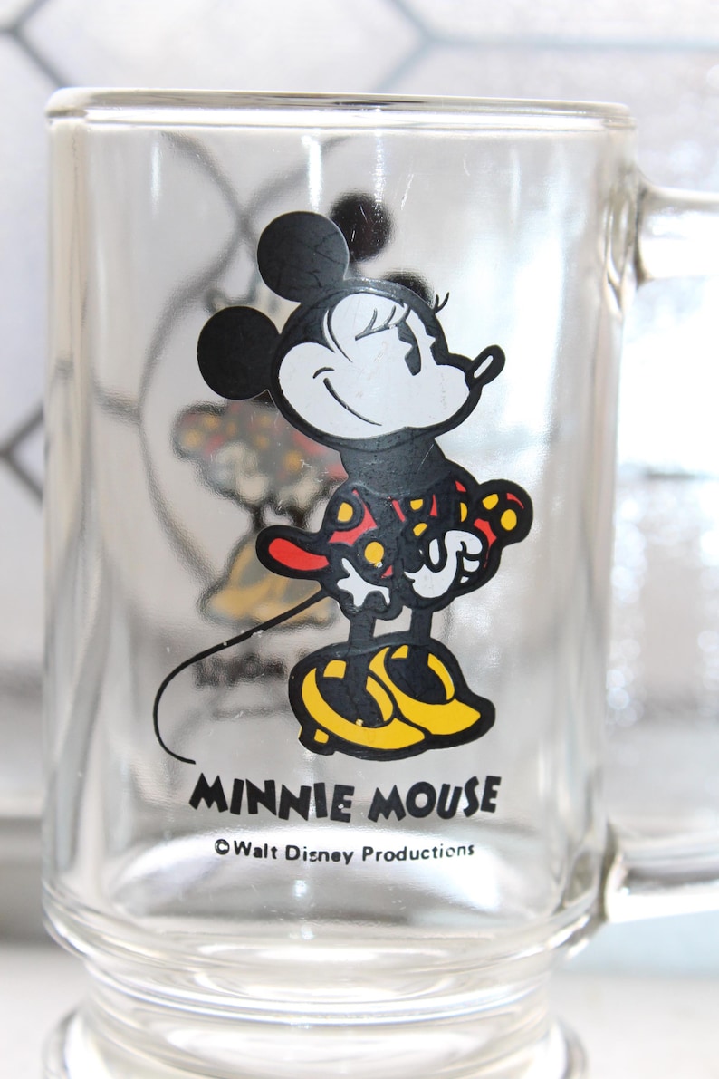 May include: A clear glass mug featuring a graphic of Minnie Mouse wearing a red dress with yellow polka dots and yellow shoes. The text MINNIE MOUSE is printed in black below the character, followed by the text &copy;Walt Disney Productions.