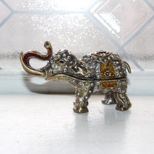 May include: A small, ornate elephant figurine made of metal with a gold finish. The elephant is decorated with rhinestones and colorful enamel. The elephant's trunk is curled up, and its eyes are inlaid with black stones.