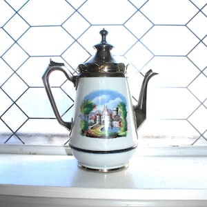 May include: White and silver vintage teapot with a scenic illustration of a castle and landscape. The teapot has a silver lid, handle, and spout. The base is white with a black band and silver trim. A decorative piece for the kitchen.