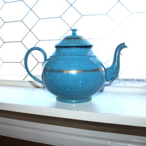 May include: A speckled turquoise teapot with a lid and handle, accented with gold bands. The teapot is sitting on a white windowsill in front of a window with a geometric pattern.