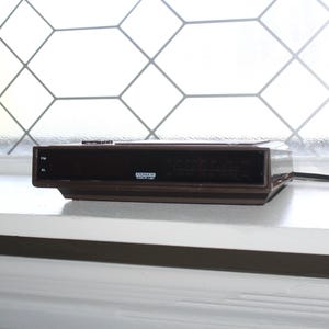 May include: A brown clock radio with a digital display and a black power cord. The clock radio is sitting on a white surface.