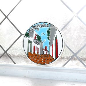 May include: A round ceramic tile featuring a hand-painted street scene with white buildings, red doors, and a blue sky. The cobblestone street and the tile's white border complete the design. The tile is displayed on a white surface.