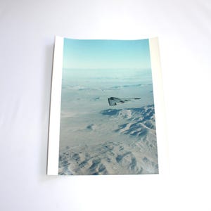 May include: A black stealth bomber aircraft flying over a desert landscape with snow-capped mountains in the distance. The sky is a light blue with white clouds.