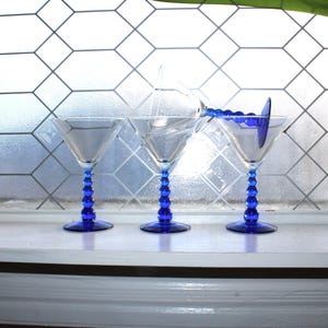 May include: Three clear martini glasses with cobalt blue stems. The stems have a beaded design. The glasses are arranged on a white windowsill in front of a window with a geometric pattern.