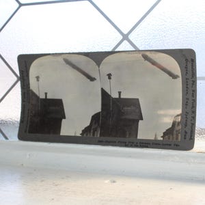 May include: Vintage stereoscopic card featuring two images of a zeppelin flying over buildings. The card is black and white, with text along the edges. The card is displayed on a white surface.