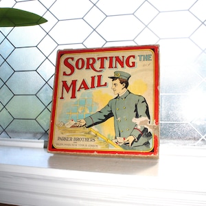 Könnte beinhalten: Vintage Parker Brothers "Sorting the Mail" Spielbox. Die Box hat einen roten Rand mit dem Titel in großen roten Buchstaben. Die Illustration zeigt einen Postboten in blauer Uniform beim Sortieren von Post. Die Box steht auf einer weißen Fensterbank.
