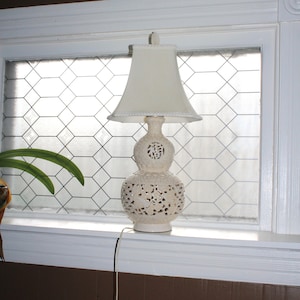 May include: A white ceramic lamp with a white lampshade sits on a windowsill. The lamp has a carved design and a round base. The window has a white grid pattern.