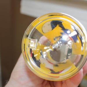 May include: A clear glass tumbler viewed from the bottom, featuring a yellow and black graphic design. The base includes embossed text that reads '12' and 'WALT DISNEY PROD. N.Y.' along with a small square moulded detail on the outer rim.