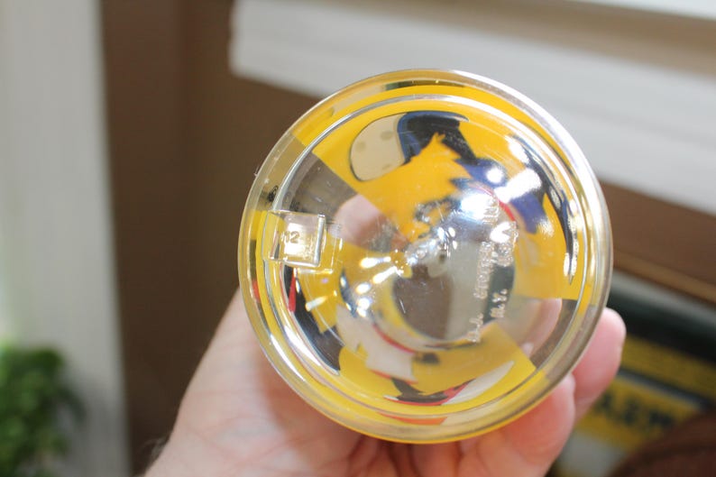 May include: A clear glass tumbler viewed from the bottom, featuring a yellow and black graphic design. The base includes embossed text that reads '12' and 'WALT DISNEY PROD. N.Y.' along with a small square moulded detail on the outer rim.