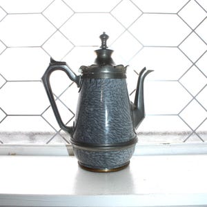 May include: A vintage gray and white enamelware teapot with a metal handle, spout, and lid. The teapot has a unique speckled pattern and sits on a white windowsill. The background features a geometric window design.