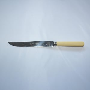 Könnte beinhalten: Ein silbernes Kuchmesser mit weißem Griff. Das Messer hat die Worte "Cake Knife" in die Klinge graviert.