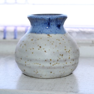 Pode incluir: Um pequeno vaso de cerâmica branca com uma borda azul. O vaso tem um esmalte salpicado e uma forma ligeiramente arredondada.